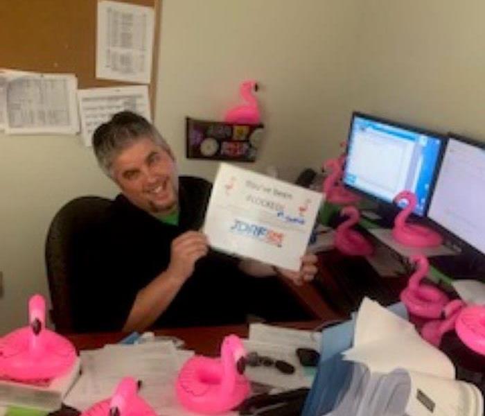 man at desk with plastic pink flamigoes all around him holding a sign saying you've been flocked